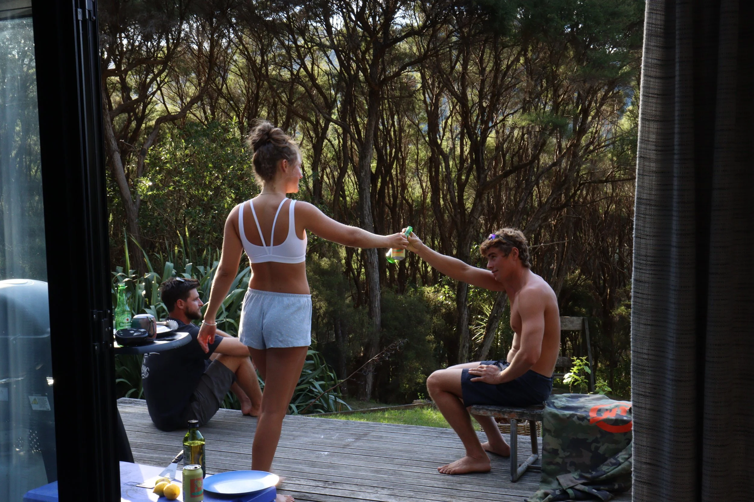 People gathering outdoors on a wooden deck with a background of trees. One woman in a white tank top and shorts is standing and holding a spray bottle, while a shirtless man is kneeling in front of her. Two other men are seated; one with a beard and dark clothing sitting on the deck, and another with glasses behind the woman. There are bottles, cups, and food items on the deck table.