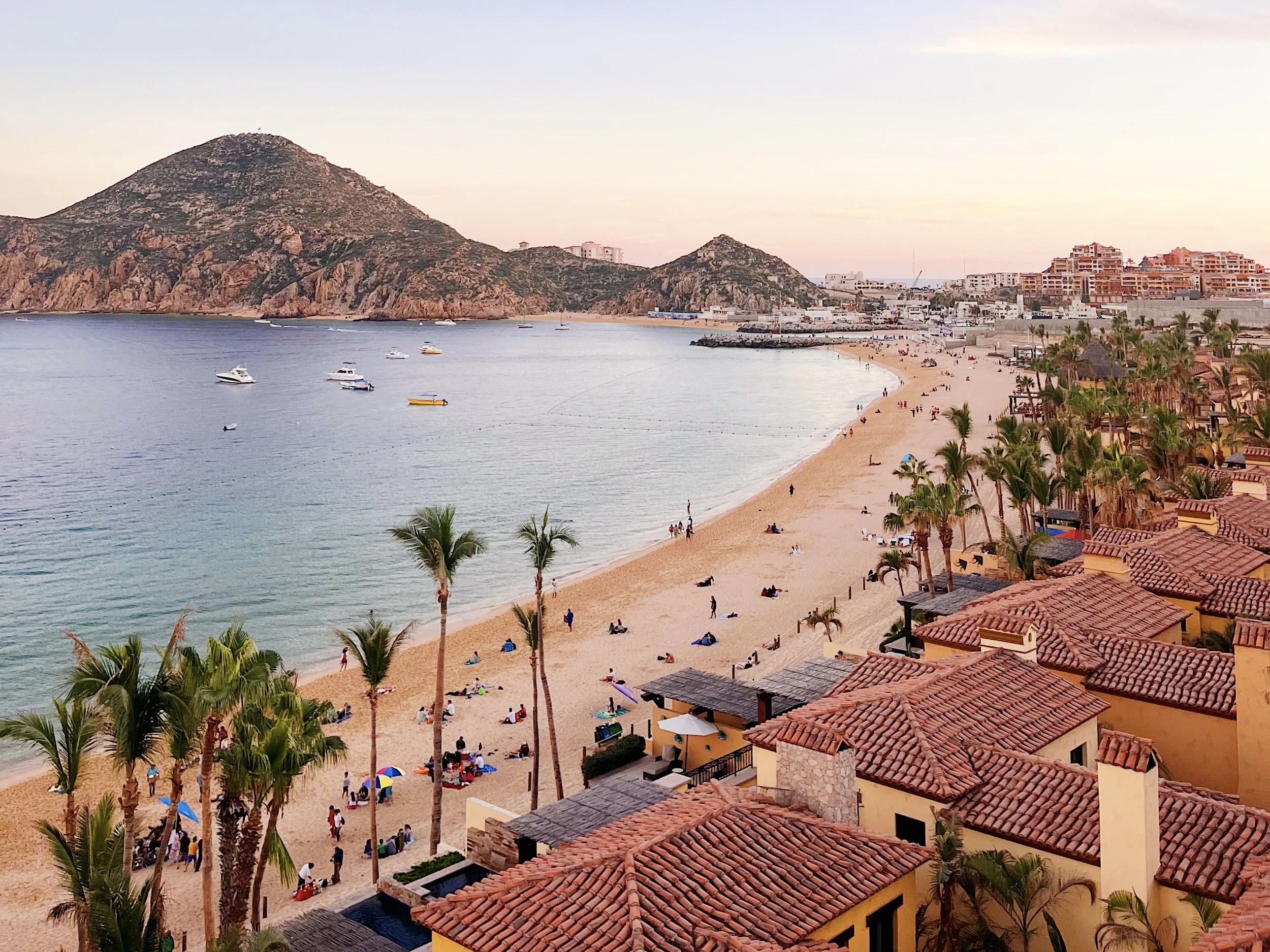 A coastal scene with a sandy beach filled with people, palm trees, and colorful umbrellas in the foreground. Several boats are floating on calm water near the shore. In the background, there are rocky hills and a mix of modern buildings and houses on the hillside, under a pastel-colored sky.