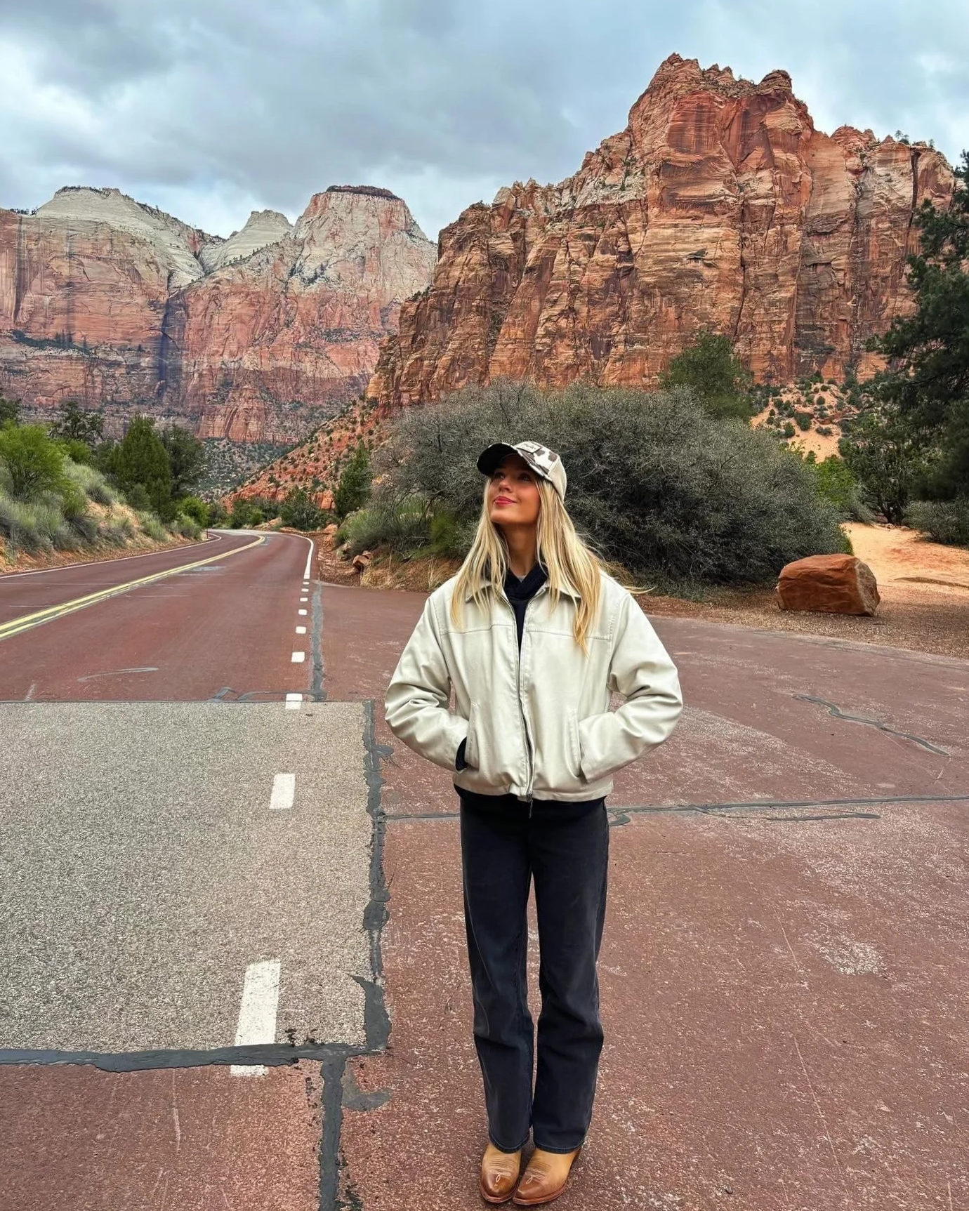 A woman with long blonde hair standing on a mountain road in front of red rock formations, wearing a beige jacket, black pants, brown shoes, and a light-colored cap, with her hands in her pockets and looking up.
