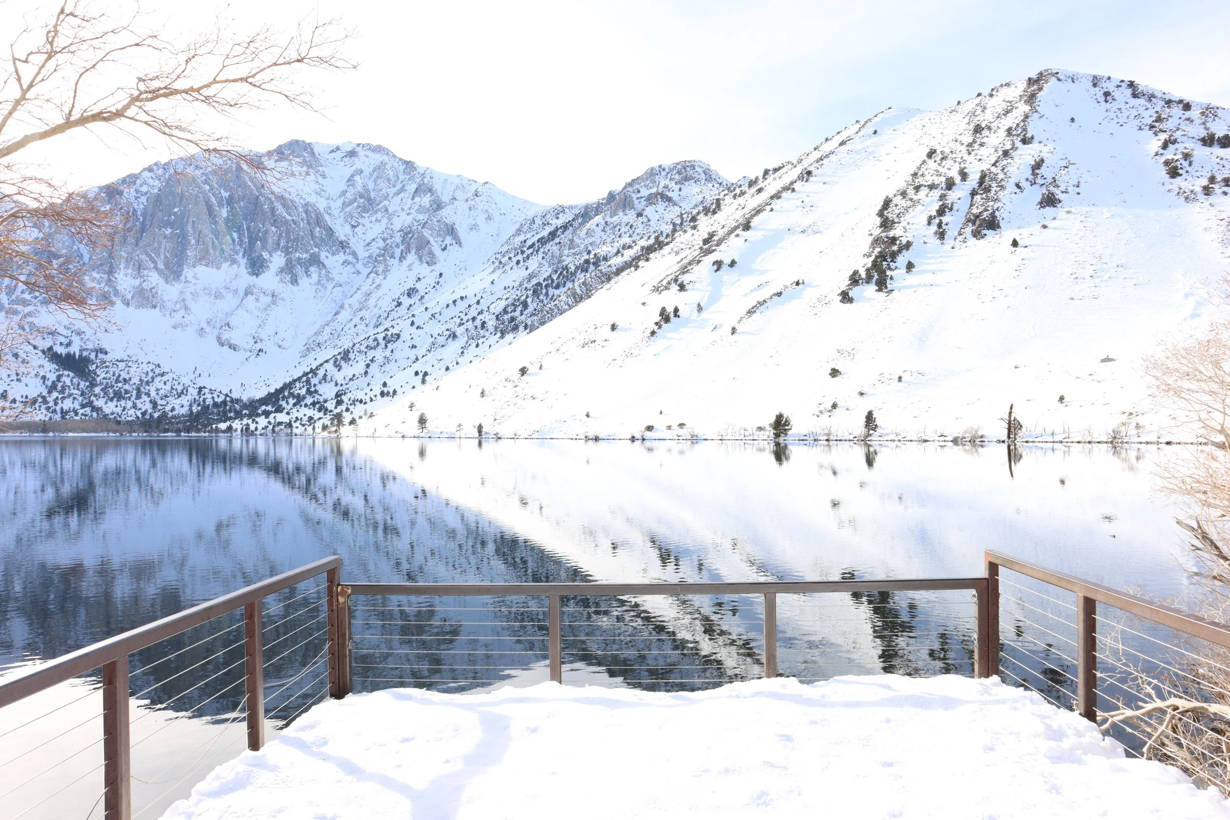 Snow-covered mountain range reflected in a calm lake, seen from a wooden deck with metal railing.