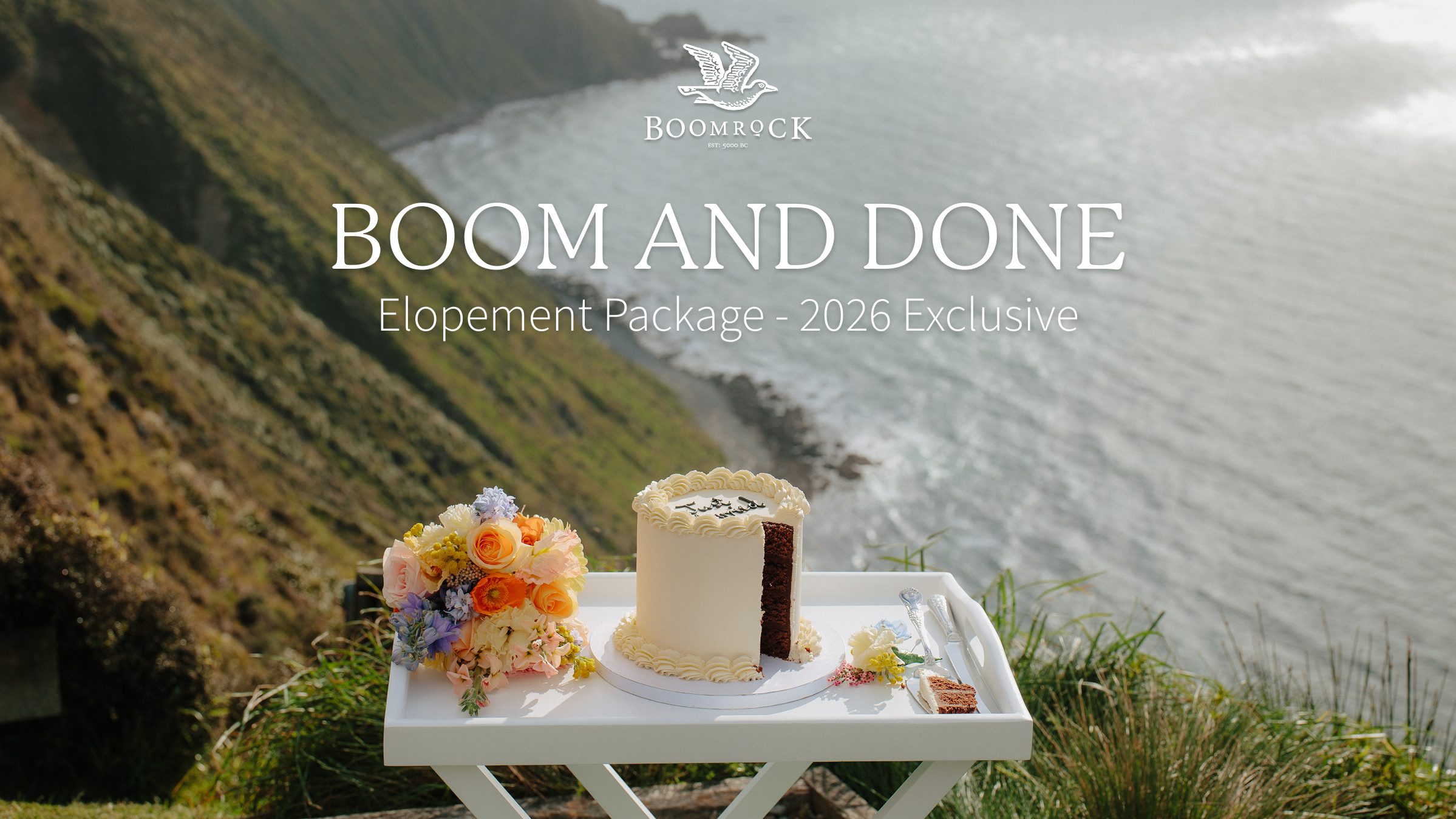 A white table with a cake, flowers, and utensils positioned outdoors on a grassy area overlooking the ocean, with cliffs and water in the background.