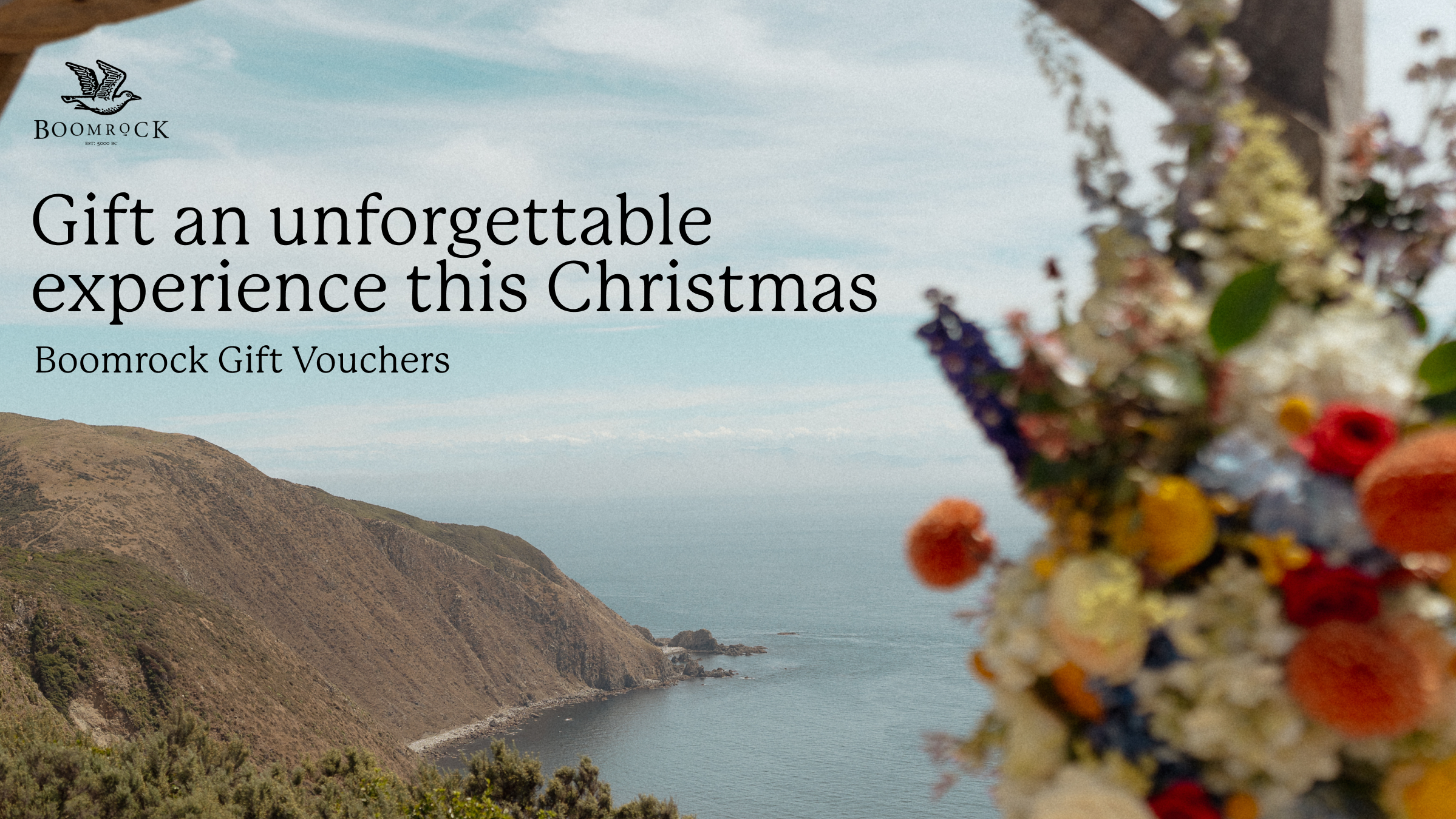 A promotional gift voucher for Boomrock, with the tagline 'Gift an unforgettable experience this Christmas.' The background features a scenic view of coastal cliffs and ocean, with a blurred foreground of colorful flowers or decorations.
