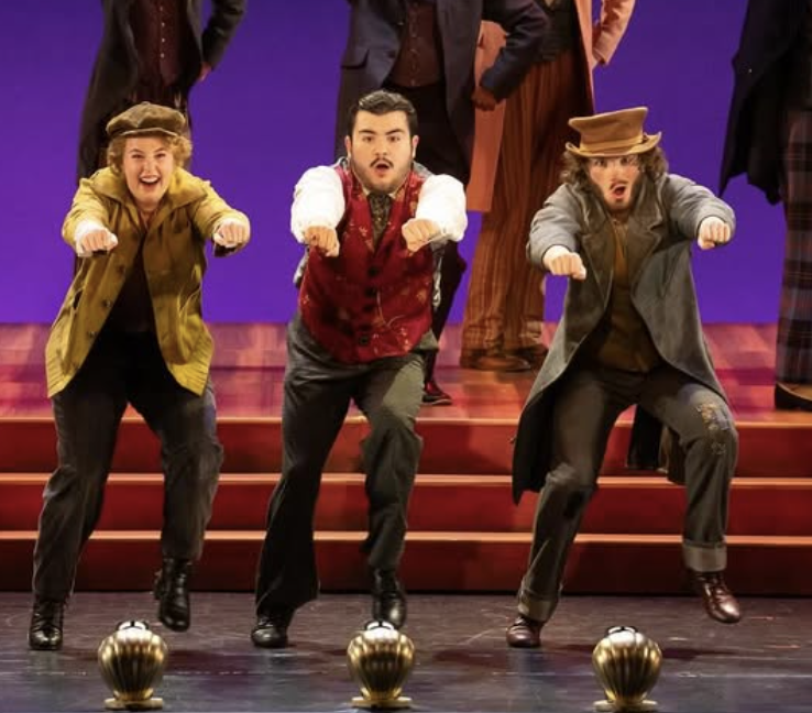 Three performers on stage wearing costumes and hats, dancing in front of a purple background with a group of people behind them. The stage has a red carpeted riser with gold balls in front.