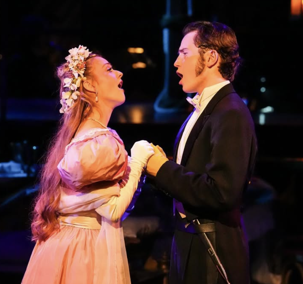 Two performers, a woman with long red hair wearing a pink dress and floral headpiece, and a man with brown hair in a tuxedo, sing closely facing each other on stage.