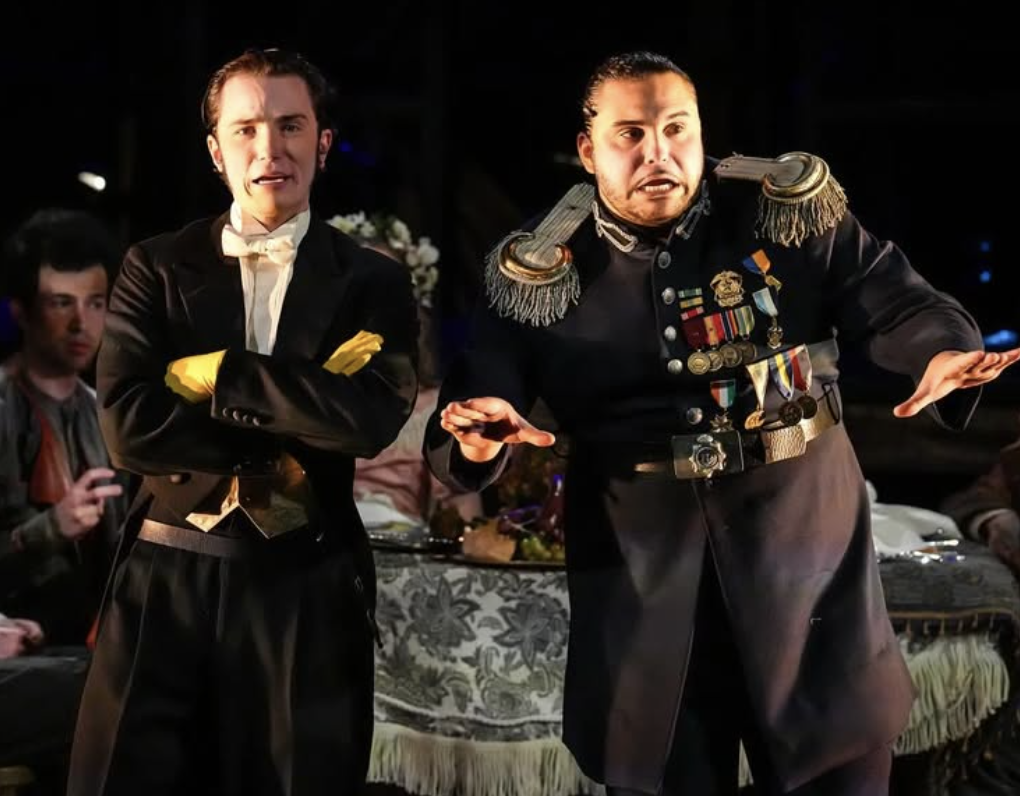 Two men in formal costumes appear to be performing on stage. One man is dressed as a military officer with medals, and the other is dressed as a man in formal evening wear, possibly a butler or a performer in a tuxedo. Both have expressive facial exp
