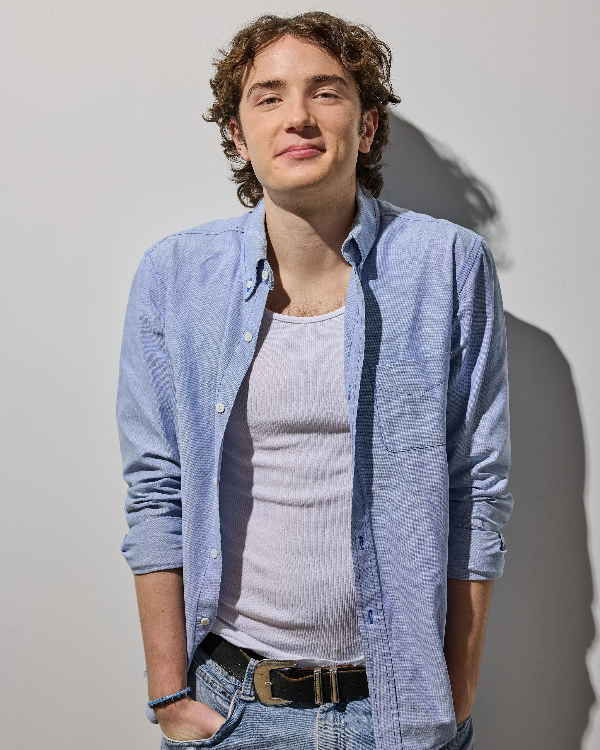 A young man with curly brown hair and fair skin wearing a light blue button-up shirt over a white tank top, with jeans, standing against a plain gray wall with his right hand in his jeans pocket.