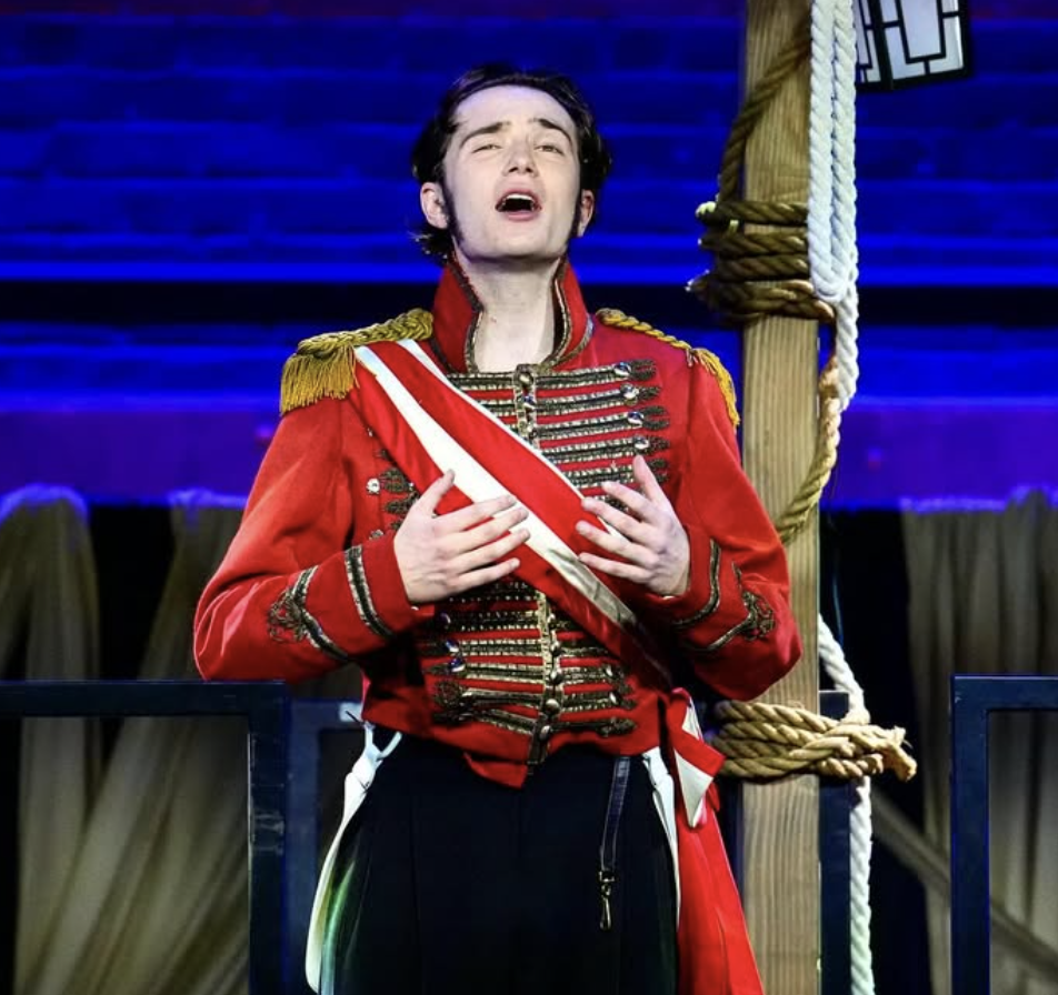 A young man dressed as a royal guard with a red jacket adorned with gold cords and shoulder epaulets, singing passionately on stage with a blue background and a rope and wooden post in the background.
