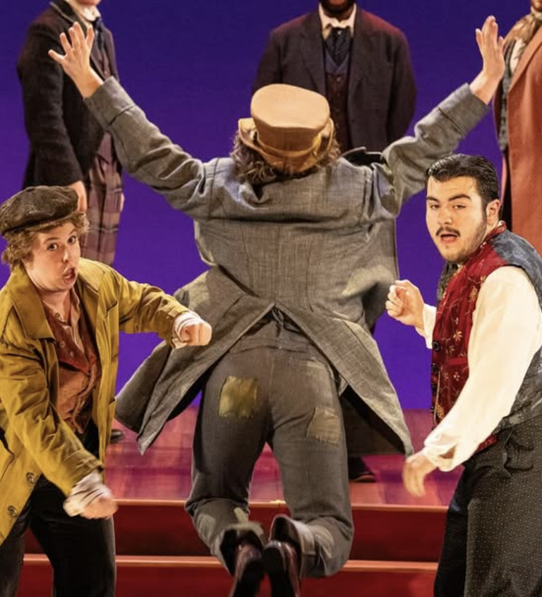Actors performing on stage, with three people in the foreground and three in the background, engaging in a theatrical scene with expressive gestures.