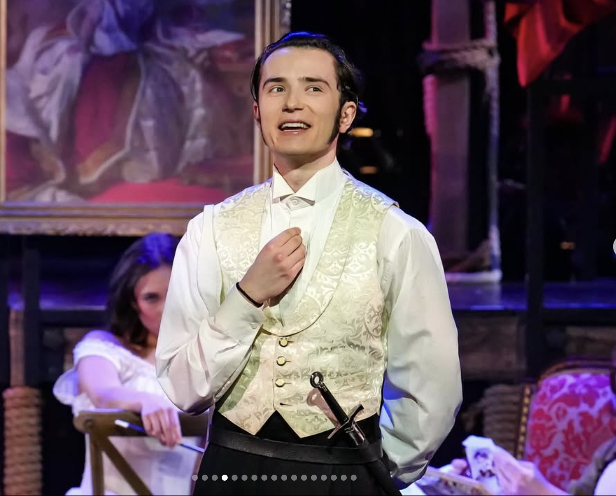 A young man with dark hair, dressed in a white shirt with a cream-colored vest, standing on stage during a theatrical performance. He appears to be singing or speaking with emotion, with a sword tucked into his waistband. In the background, there are