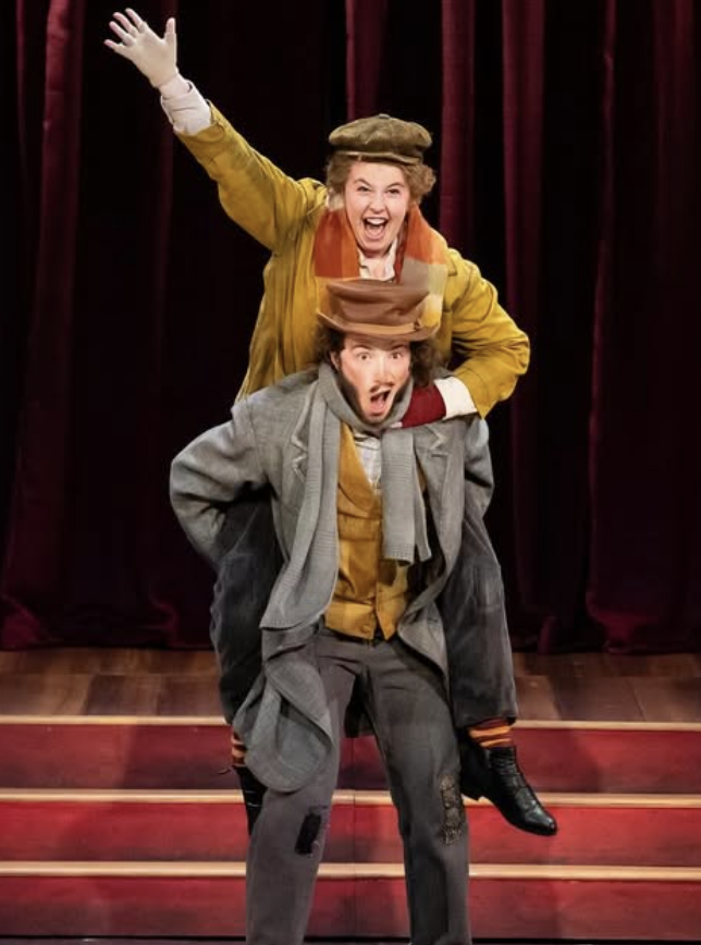 Two performers, one woman and one man, involved in a theatrical act on stage. The woman, with gray hair, is smiling and raising her arm, while the man, with a surprised expression, is dressed in vintage clothing and hats, appearing to be caught in a 
