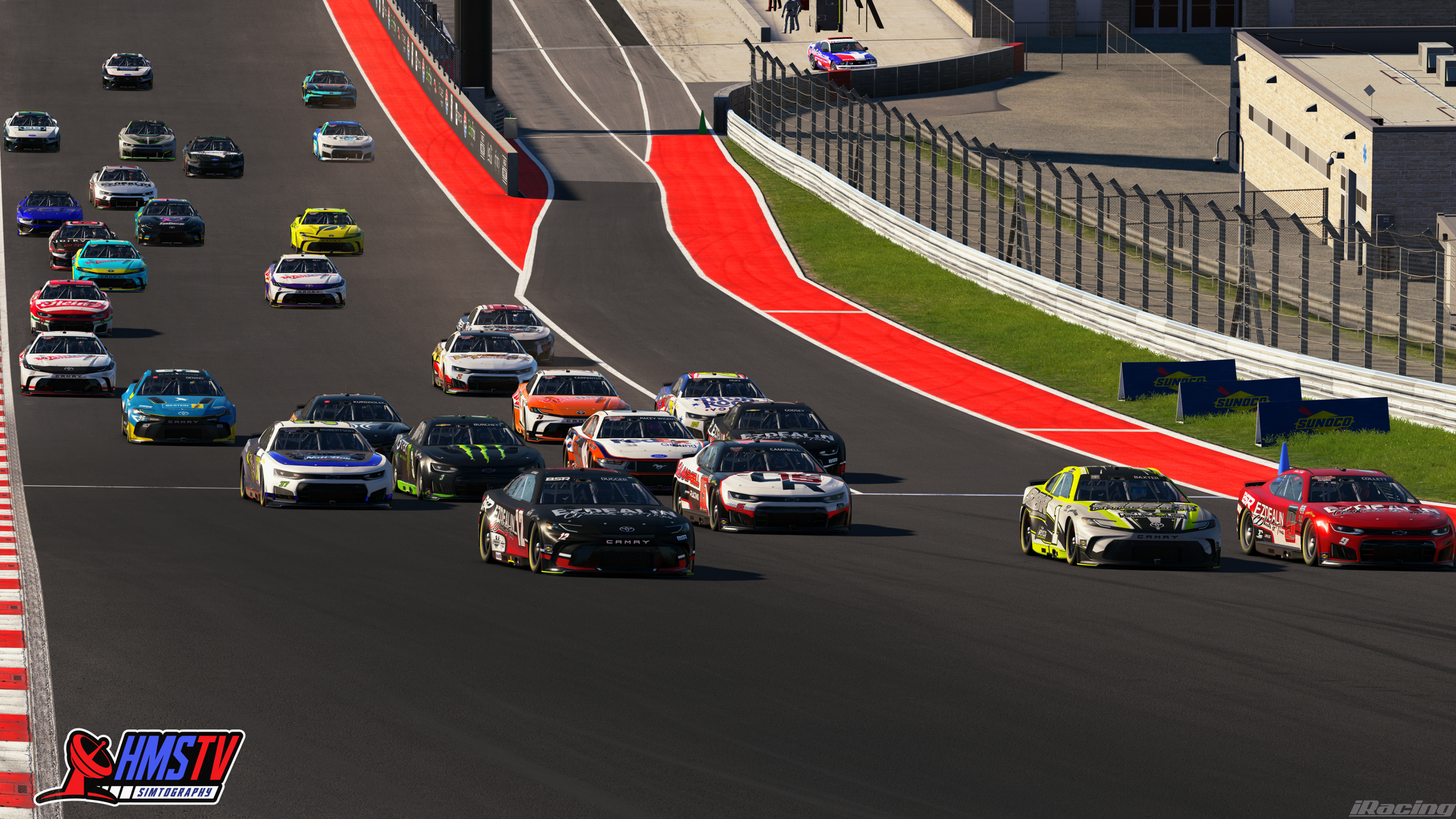 Race cars on a track in a racing event, with multiple colorful cars leading into a turn, some cars are off the racing line. Brands like Toyota and Camry are visible, with branding on the cars and trackside banners. The scene is set on a sunny day wit