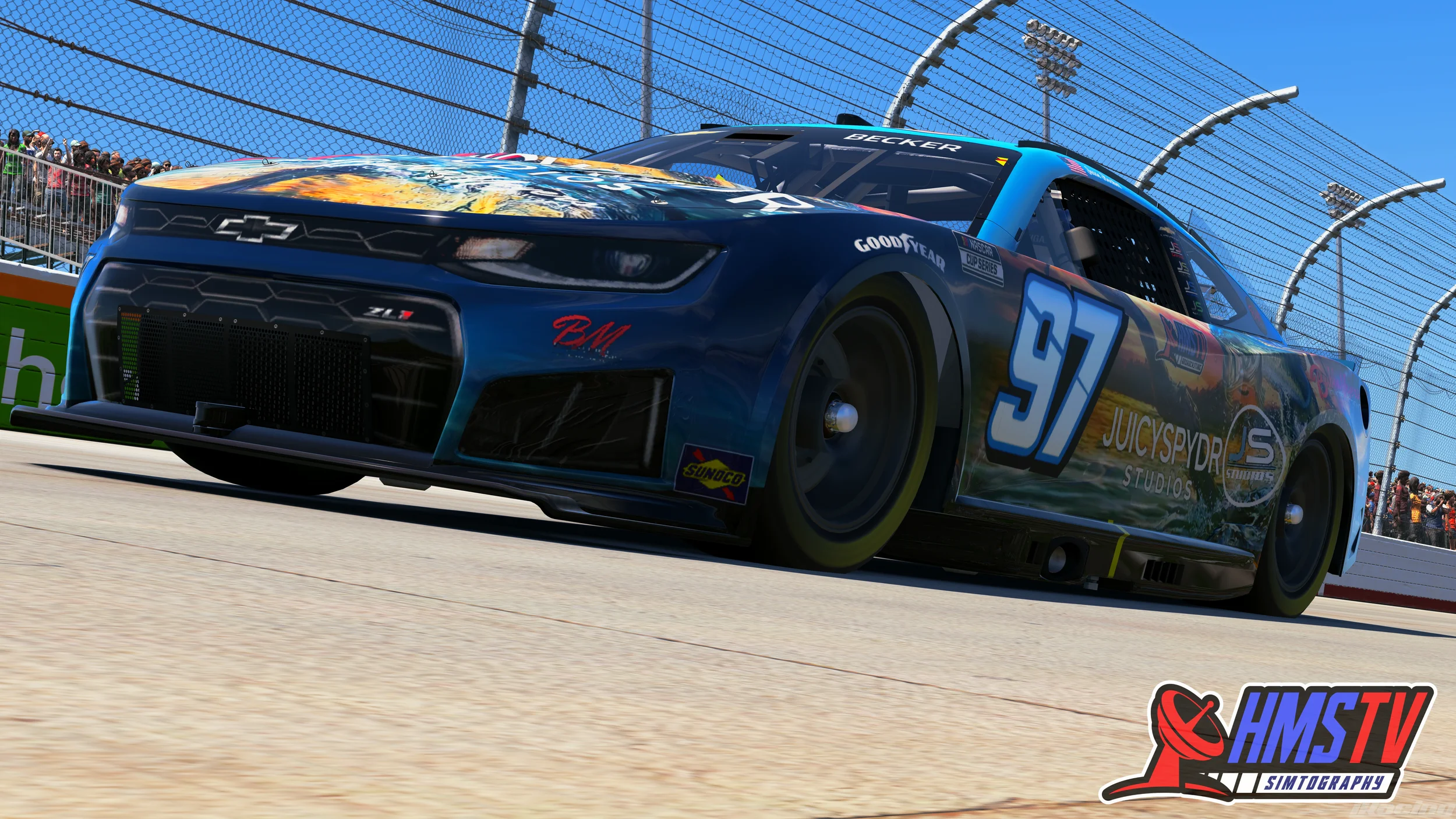 A race car with the number 97 on the side, featuring a colorful design including the logo for JucySpyder Studios, driving on a race track with spectators and fence in the background on a bright, sunny day.