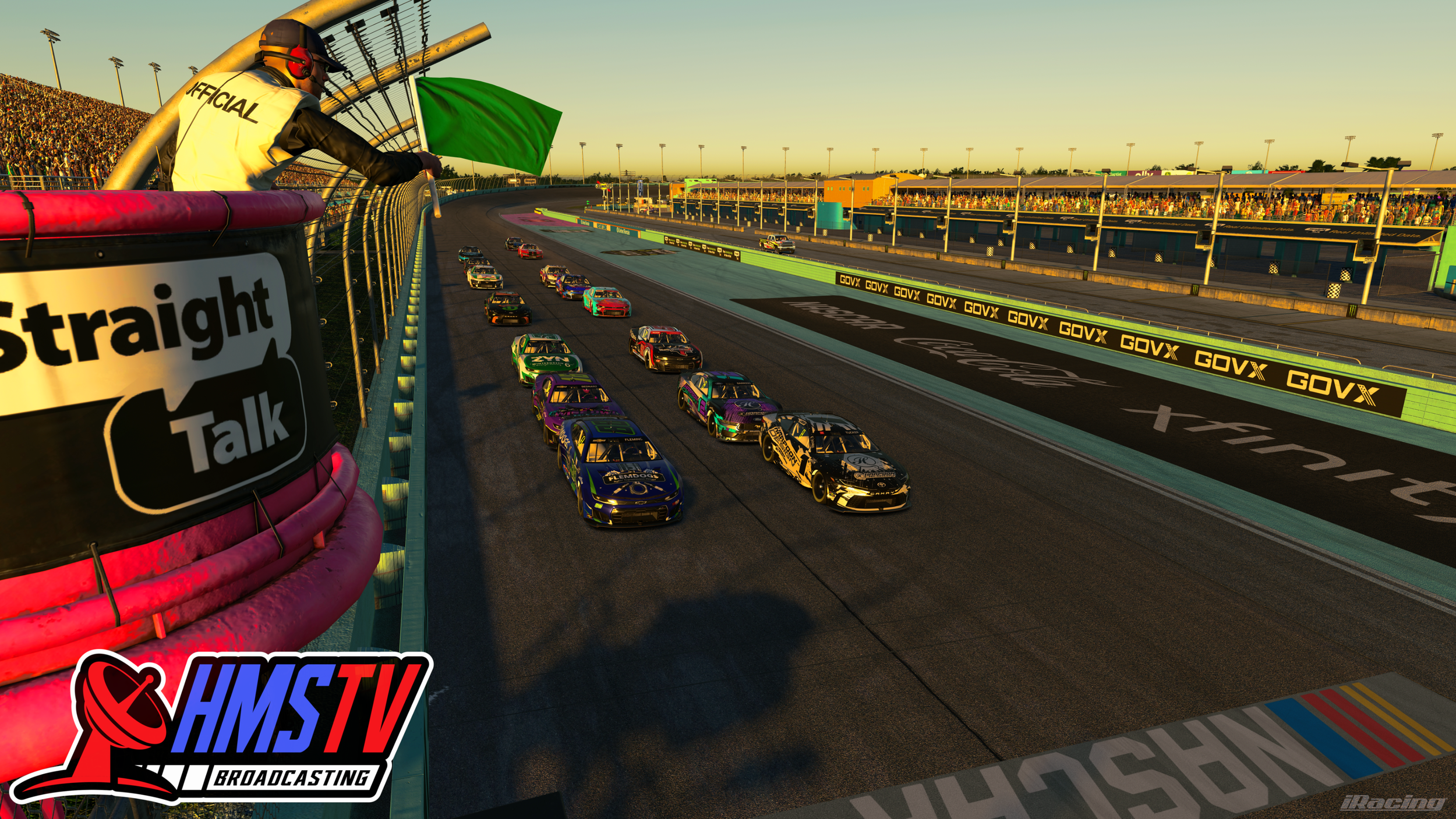 Race cars on a track with a official signaling from the pit wall, audience in the stands, and sunset lighting.