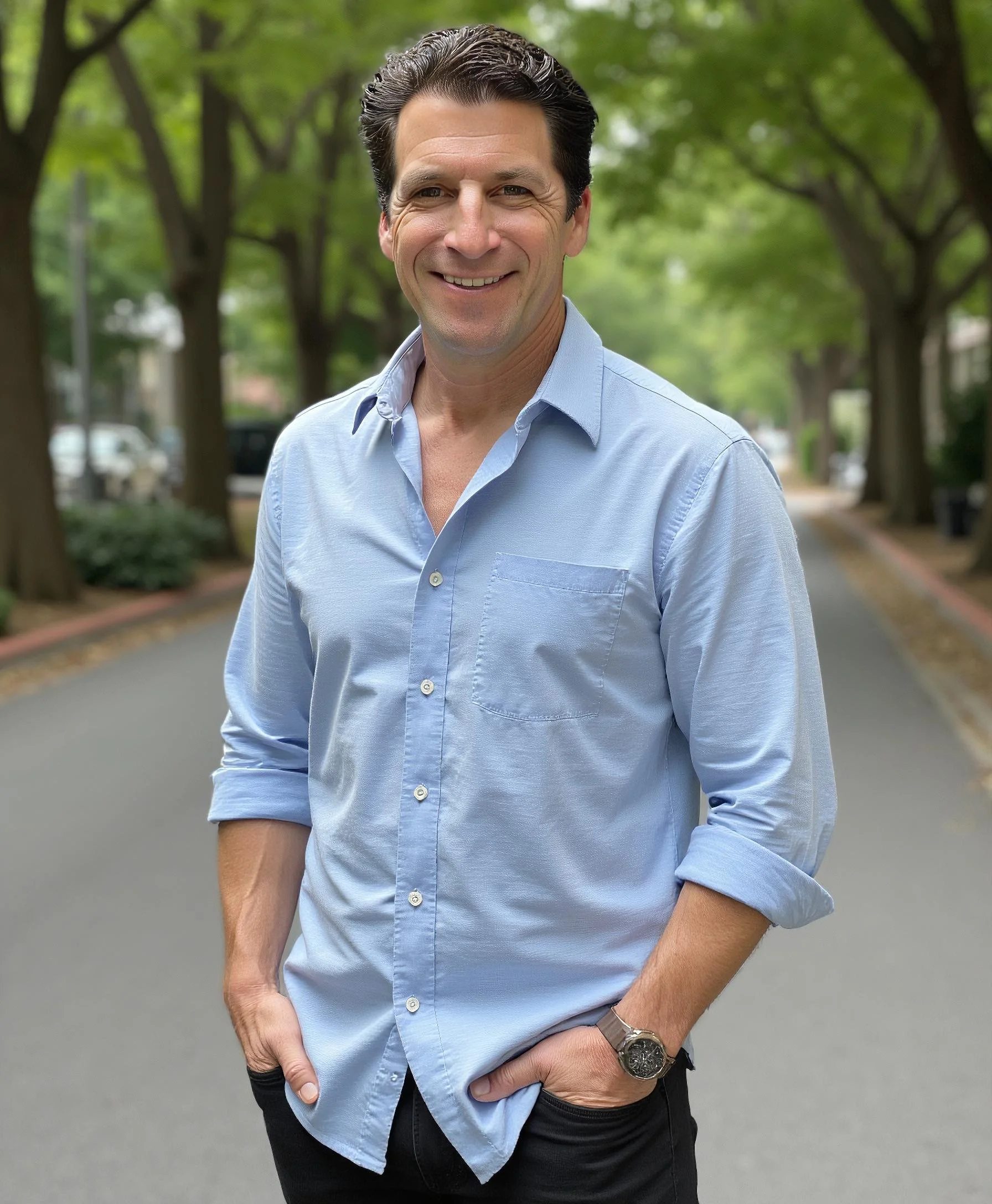 A smiling man with dark, slicked-back hair wearing a light blue button-down shirt with the sleeves rolled up. He is standing outdoors on a tree-lined street, with parked cars visible in the background.