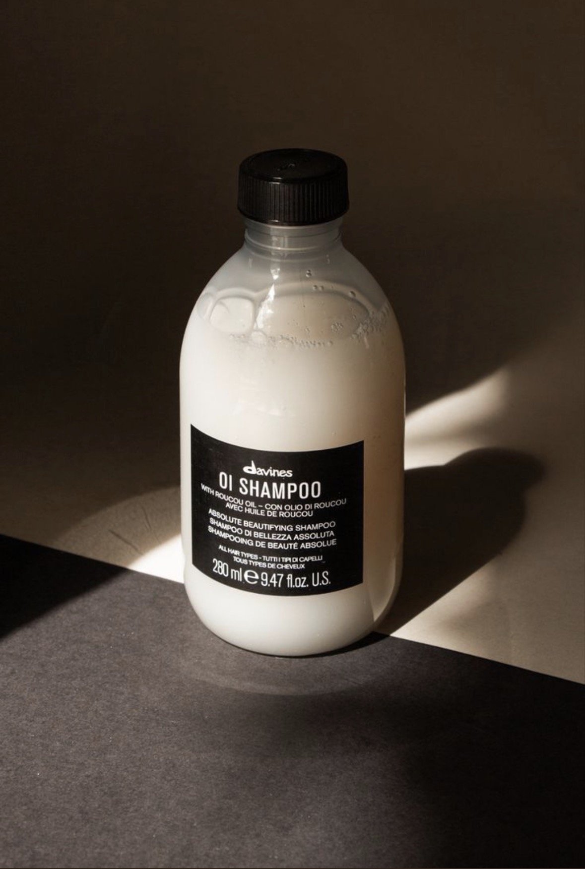A bottle of Davines OI Shampoo with Roucou oil on a dark surface with a shadow cast beside it.