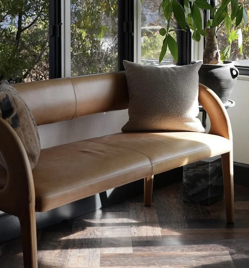 A tan leather sofa with a large gray cushion, sitting in front of windows with greenery outside and a potted plant on an adjacent table.