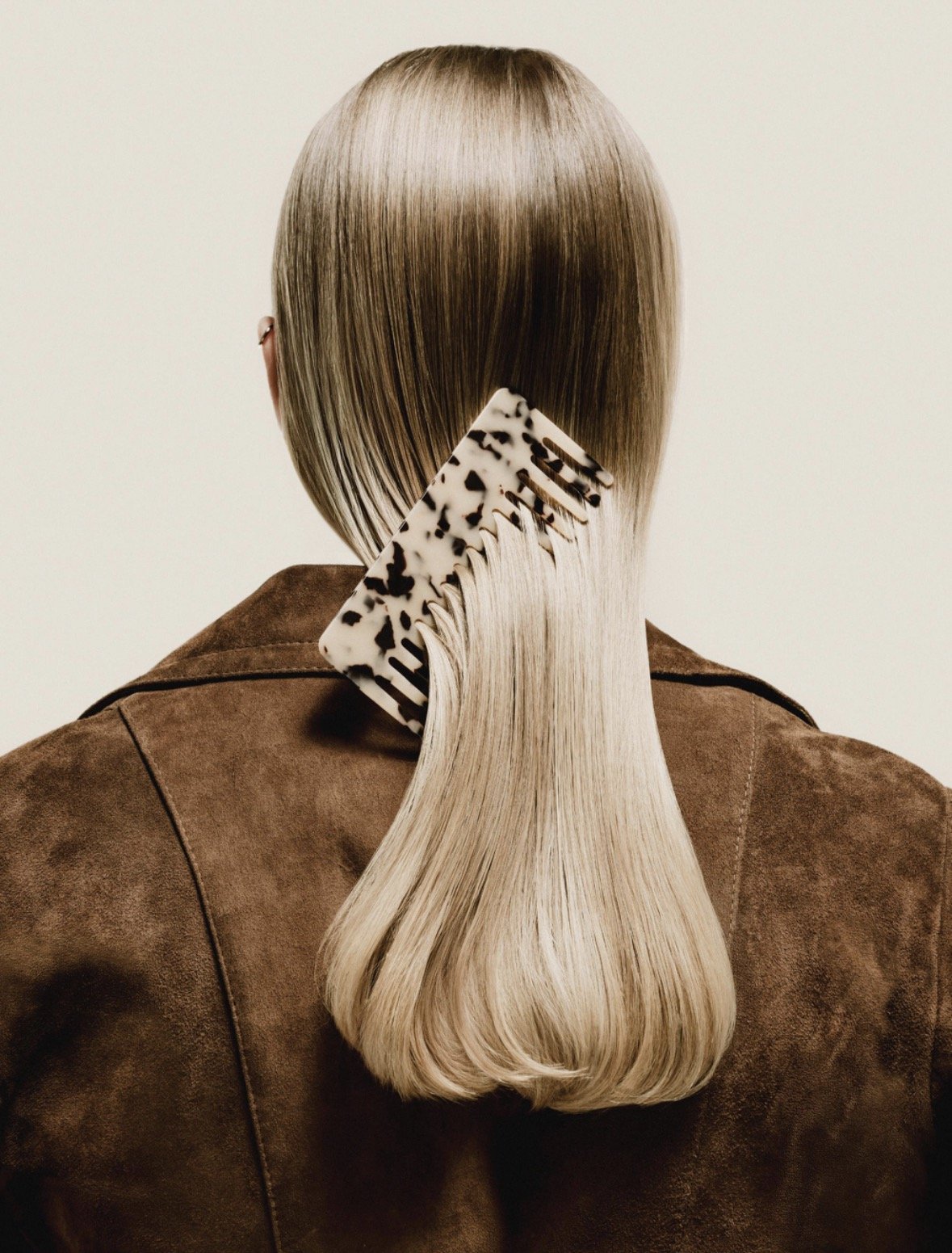Back view of a person with shiny, straight blonde hair combed with a white and black tortoiseshell hairbrush, wearing a brown suede jacket.