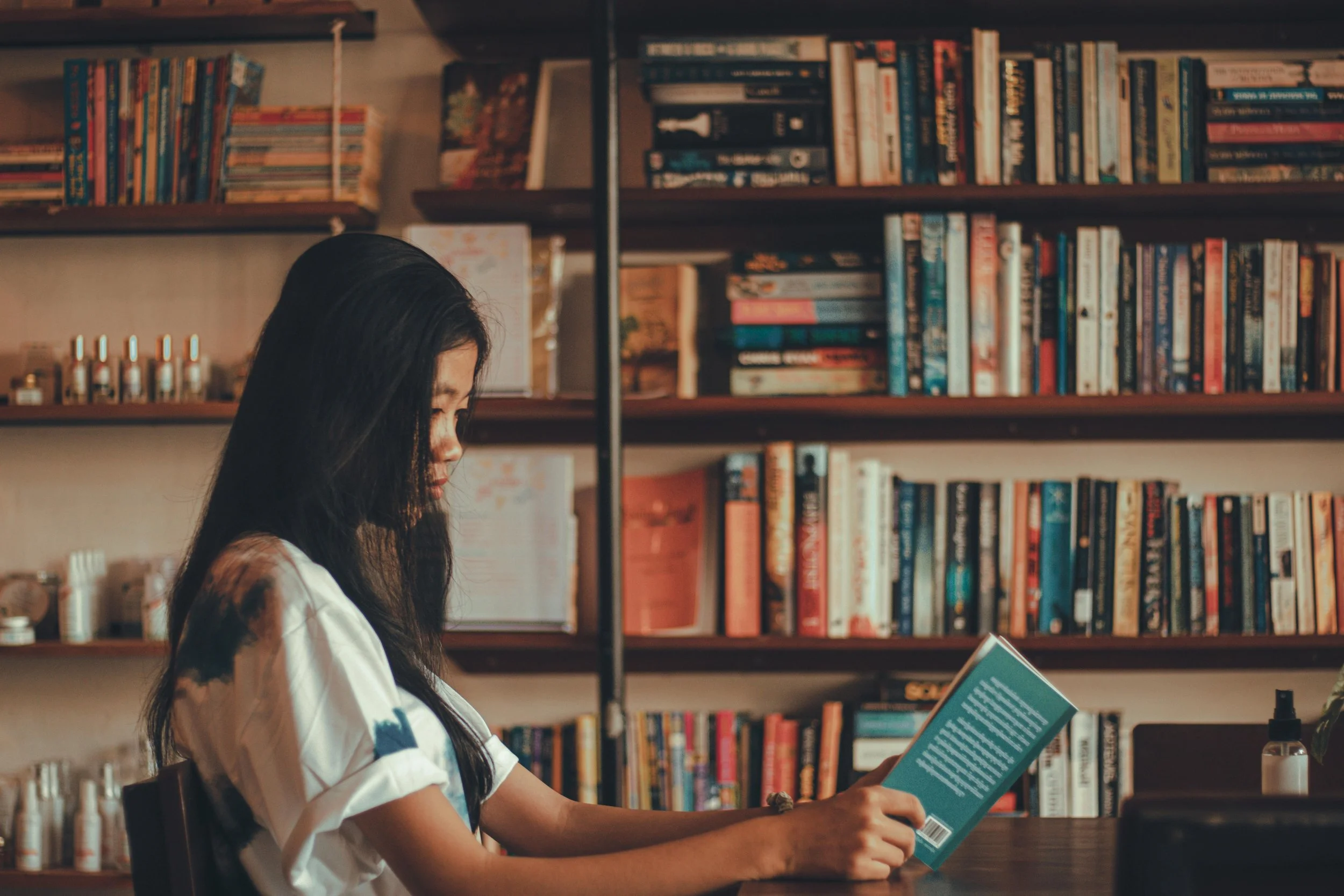 A woman with long black hair is sitting at a table in a bookstore or library, reading a book with a blue cover, with bookshelves filled with numerous books behind her.