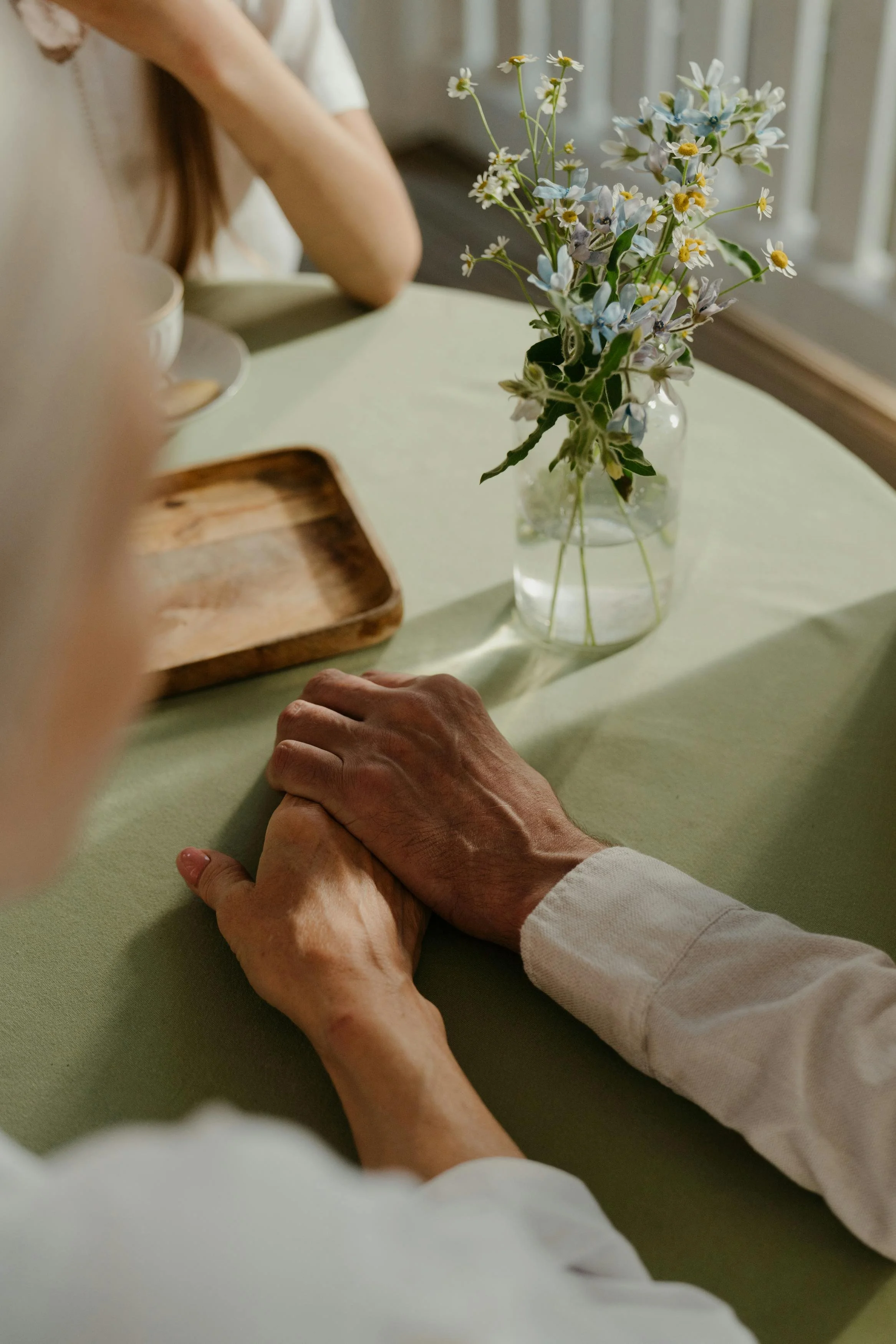 Close-up of elderly hands holding each other, resting on a green table, with a small vase of flowers and a wooden tray nearby.