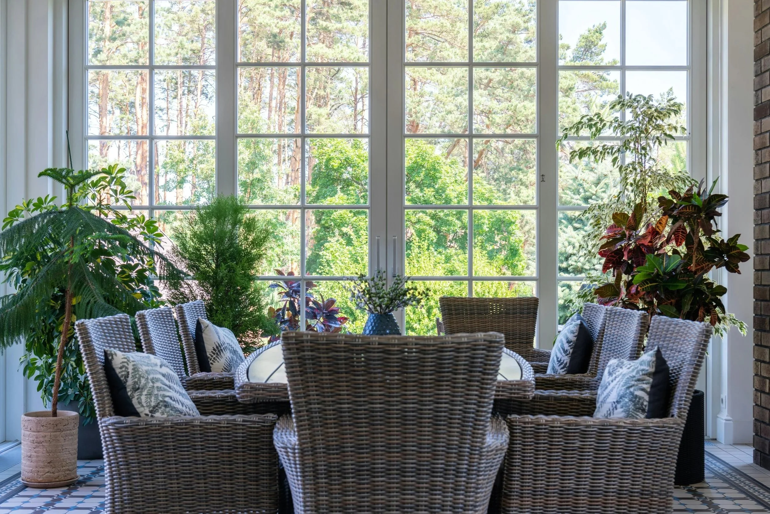 Indoor dining area with a large window view of trees outside. Wicker chairs with patterned cushions around a table with a floral centerpiece, potted plants, and a brick wall on the right.