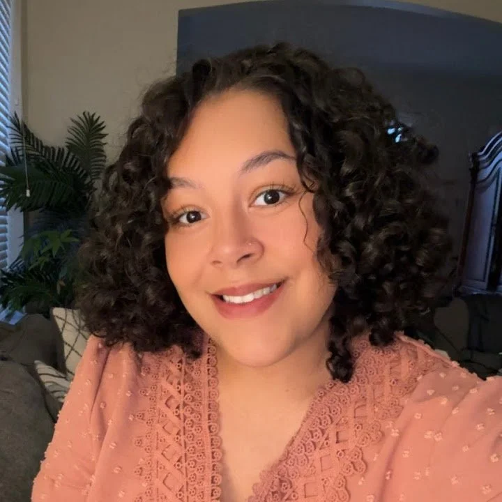 A young woman with curly hair smiling indoors, wearing a pink top with lace details, in a room with a gray headboard, patterned pillows, and a houseplant in the background.