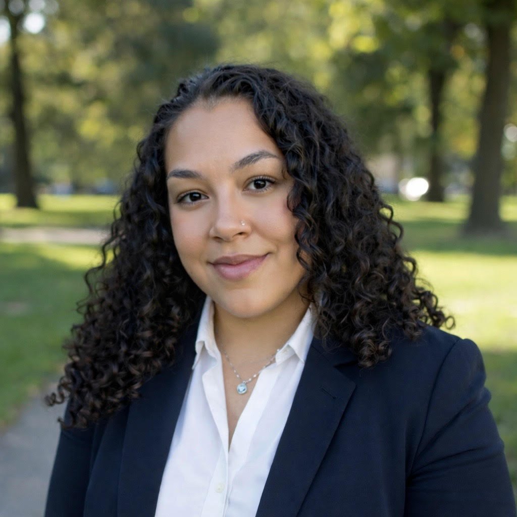 A woman with curly dark hair, wearing a navy blazer and white shirt, standing outdoors with trees and a grassy park behind her.
