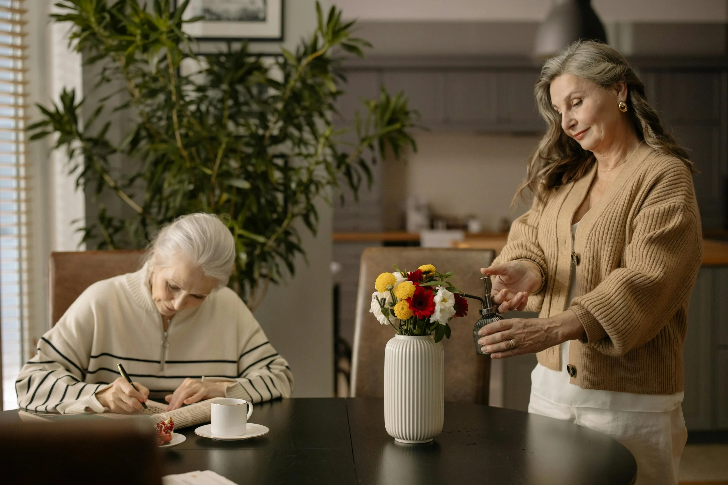 An elderly woman is sitting at a table writing in a notebook, with a coffee mug nearby. Another woman stands next to her, preparing a flower arrangement in a tall, white vase. The room has a large plant and picture frames on the wall, and is filled with natural light.