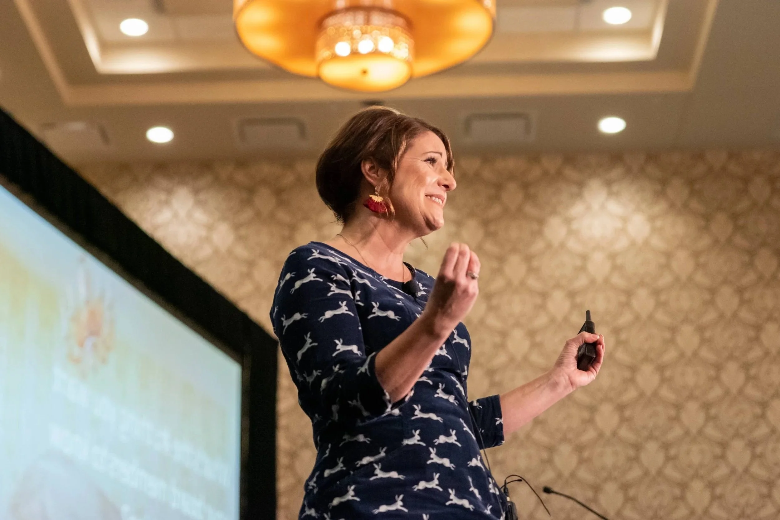 Rachel Muir, dressed in a playful navy dress adorned with white bunnies, passionately engages her audience during a vibrant fundraising workshop, radiating warmth and humor under golden lighting.
