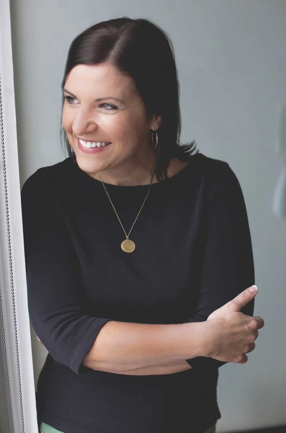 Rachel Muir, a warm and approachable nonprofit fundraising consultant, smiles brightly, wearing a black top and gold necklace, exuding generosity and fun against a soft, light background.