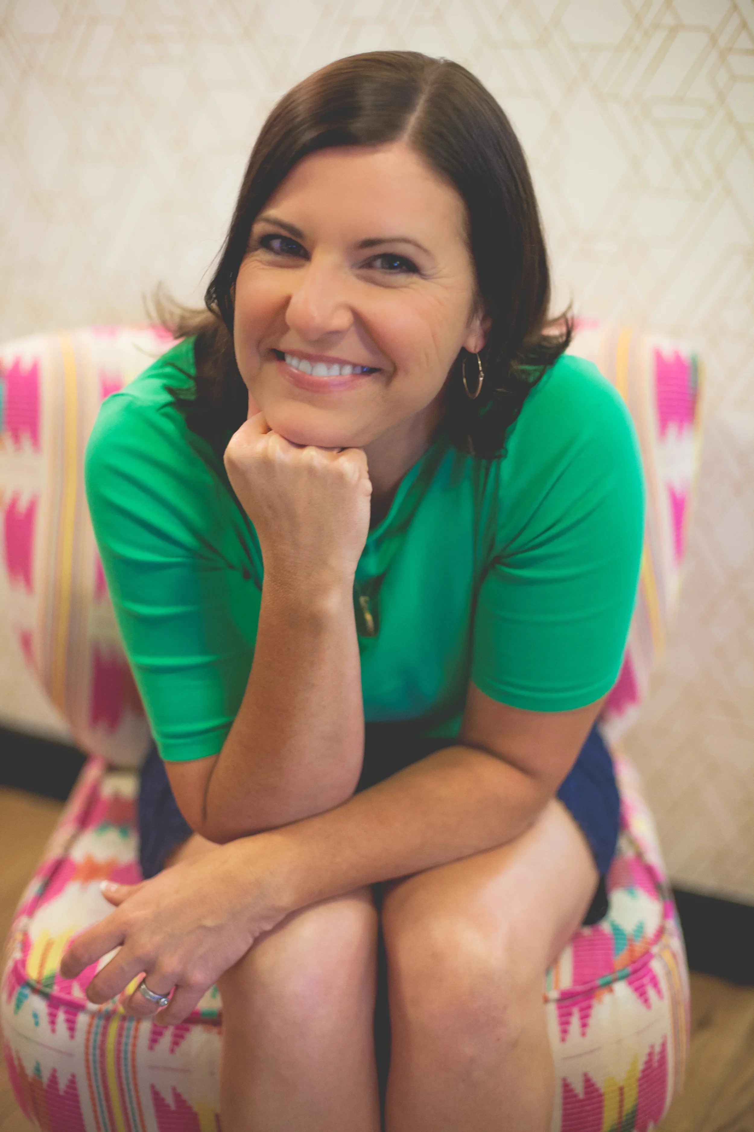 Rachel Muir, a cheerful nonprofit fundraising consultant, relaxes on a vibrant, patterned chair, wearing a bright green top and radiating warmth, ready to make fundraising fun and effective.