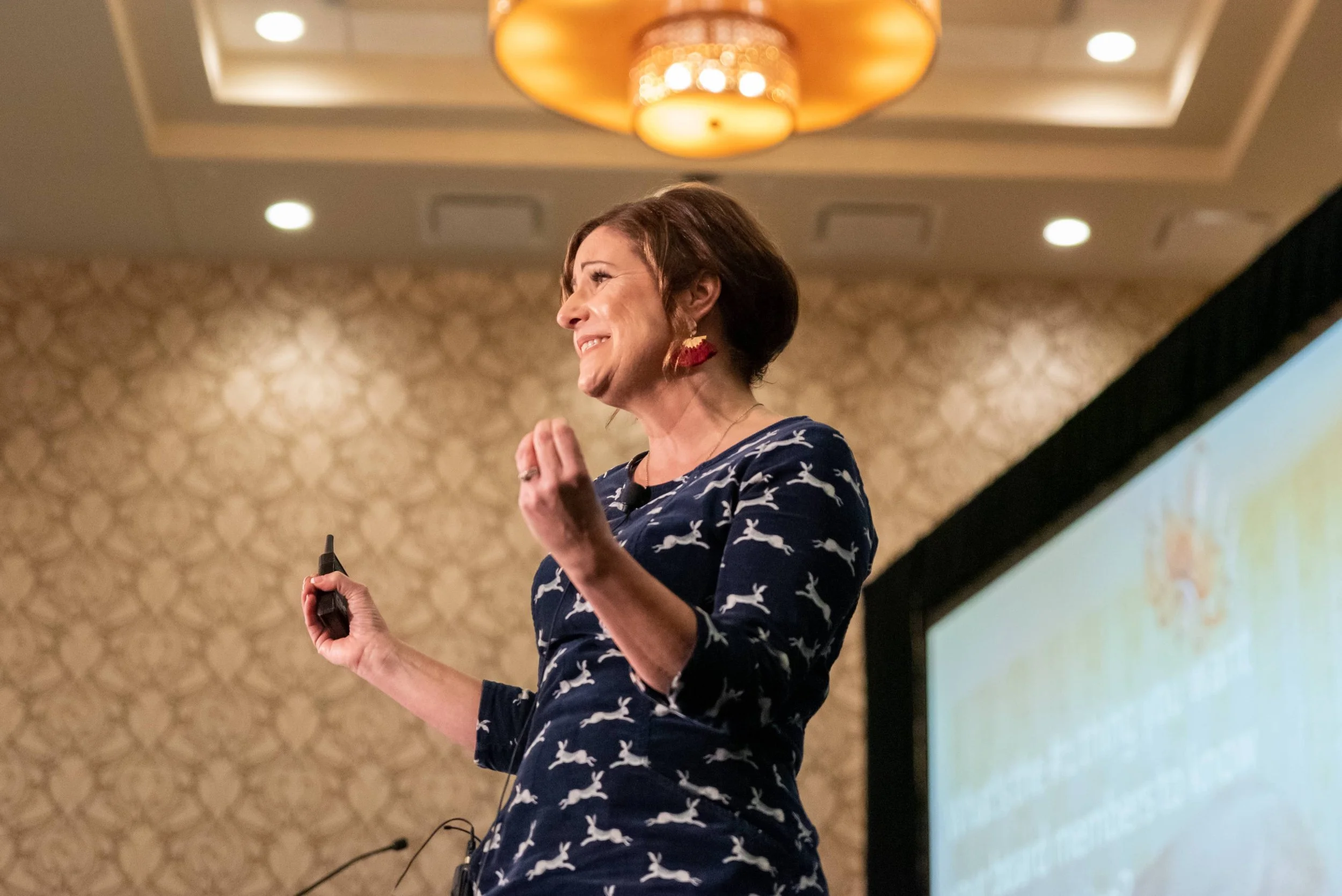 Rachel Muir, dressed in a playful navy dress adorned with white bunnies, passionately engages her audience during a vibrant fundraising workshop, radiating warmth and humor under golden lighting.