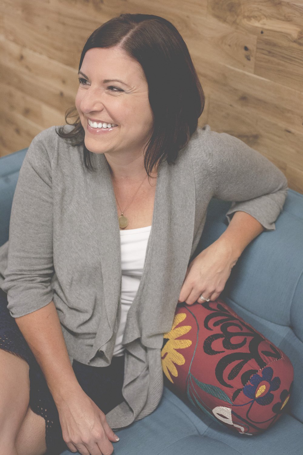 Rachel Muir, a warm and approachable nonprofit fundraising consultant, smiles brightly while seated on a teal couch, adding a pop of color with a vibrant floral pillow.