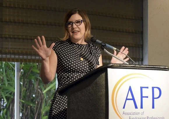 Rachel Muir, in a playful leopard print dress, passionately speaks into a microphone, engaging a captivated audience in a warm and fun atmosphere, embodying the essence of effective nonprofit fundraising.