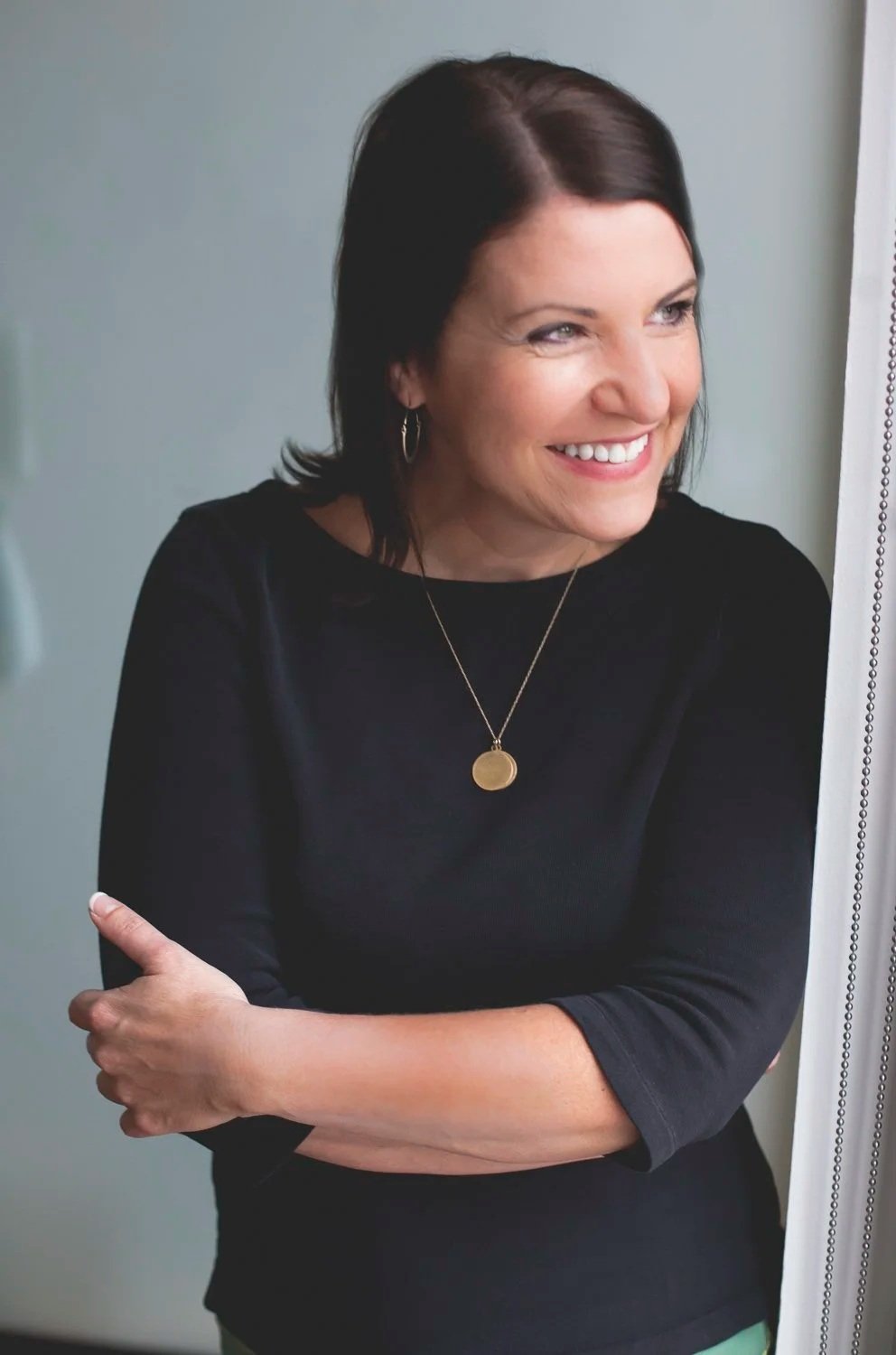 Rachel Muir, a warm and approachable nonprofit fundraising consultant, smiles brightly, wearing a black top and gold necklace, exuding generosity and fun against a soft, light background.