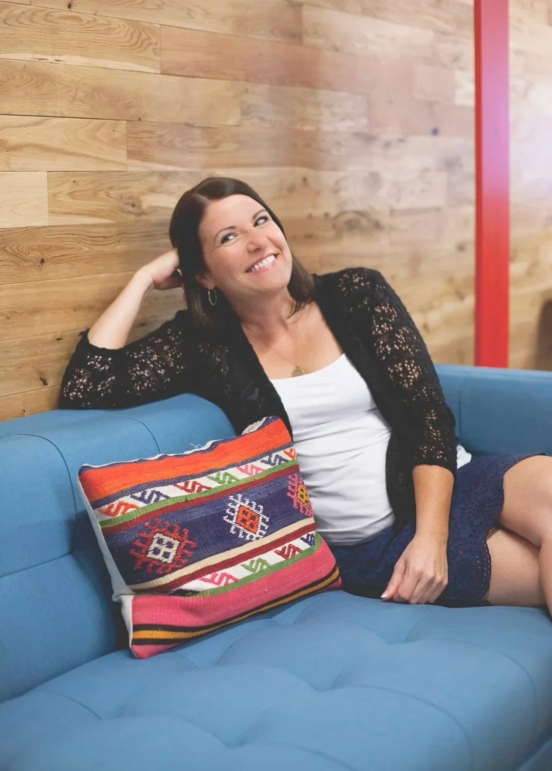 Rachel Muir smiles warmly while lounging on a vibrant blue couch, her stylish black cardigan complementing the colorful, patterned pillow beside her. A cozy, inviting space perfect for nonprofit fundraising conversations.