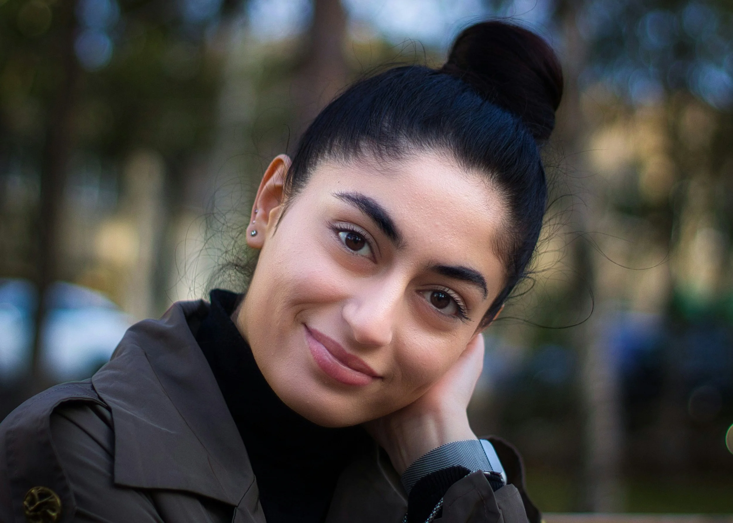 Close-up of a young woman with dark hair styled in a bun, wearing a dark jacket and a smartwatch, outdoors with blurred trees in the background.