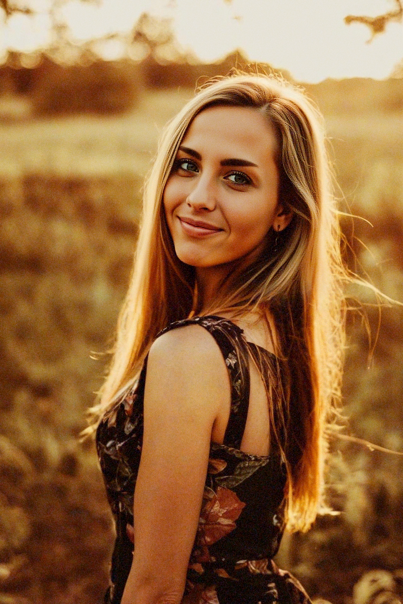 A smiling young woman with long blonde hair stands outdoors during sunset, wearing a sleeveless dress with a floral pattern.