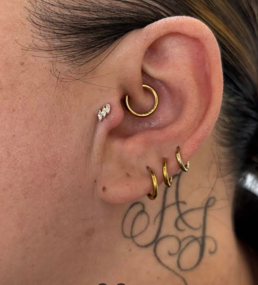 Close-up of a person's ear with multiple gold and diamond earrings and a tattoo of black cursive initials on the neck.