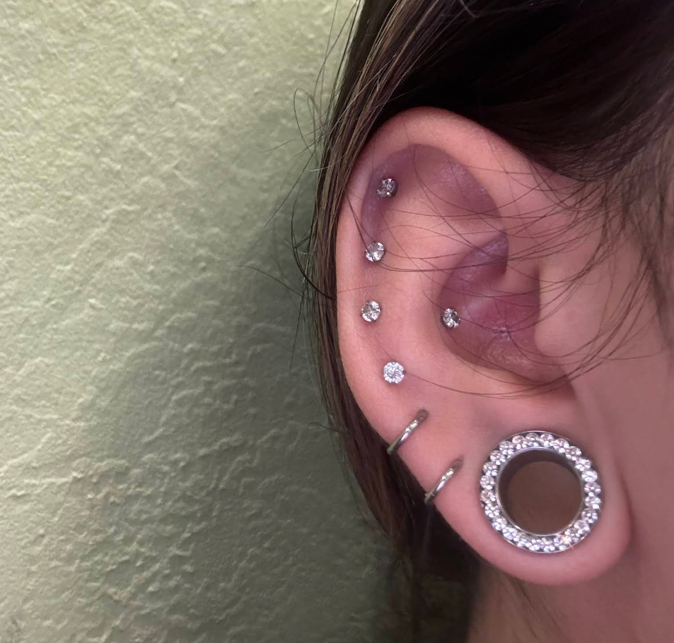 A close-up of a person's ear with multiple piercings and jewelry, including small stud earrings, a large hoop earring with rhinestones, and two thin hoops.