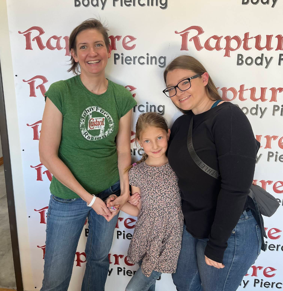 Three smiling females posing together in front of a banner that says "RapTure Body Piercing". The woman on the left is wearing a green T-shirt and jeans, the girl in the middle has a floral dress, and the woman on the right is wearing glasses, a blac