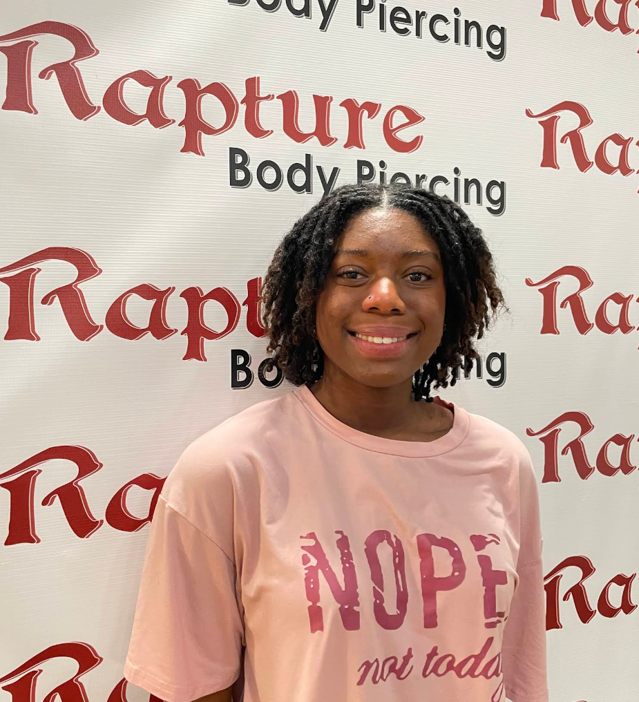 A woman with shoulder-length curly hair smiling, wearing a pink T-shirt with the words 'NOPE not today' on it, standing in front of a backdrop with the words 'Rapture Body Piercing' printed repeatedly.