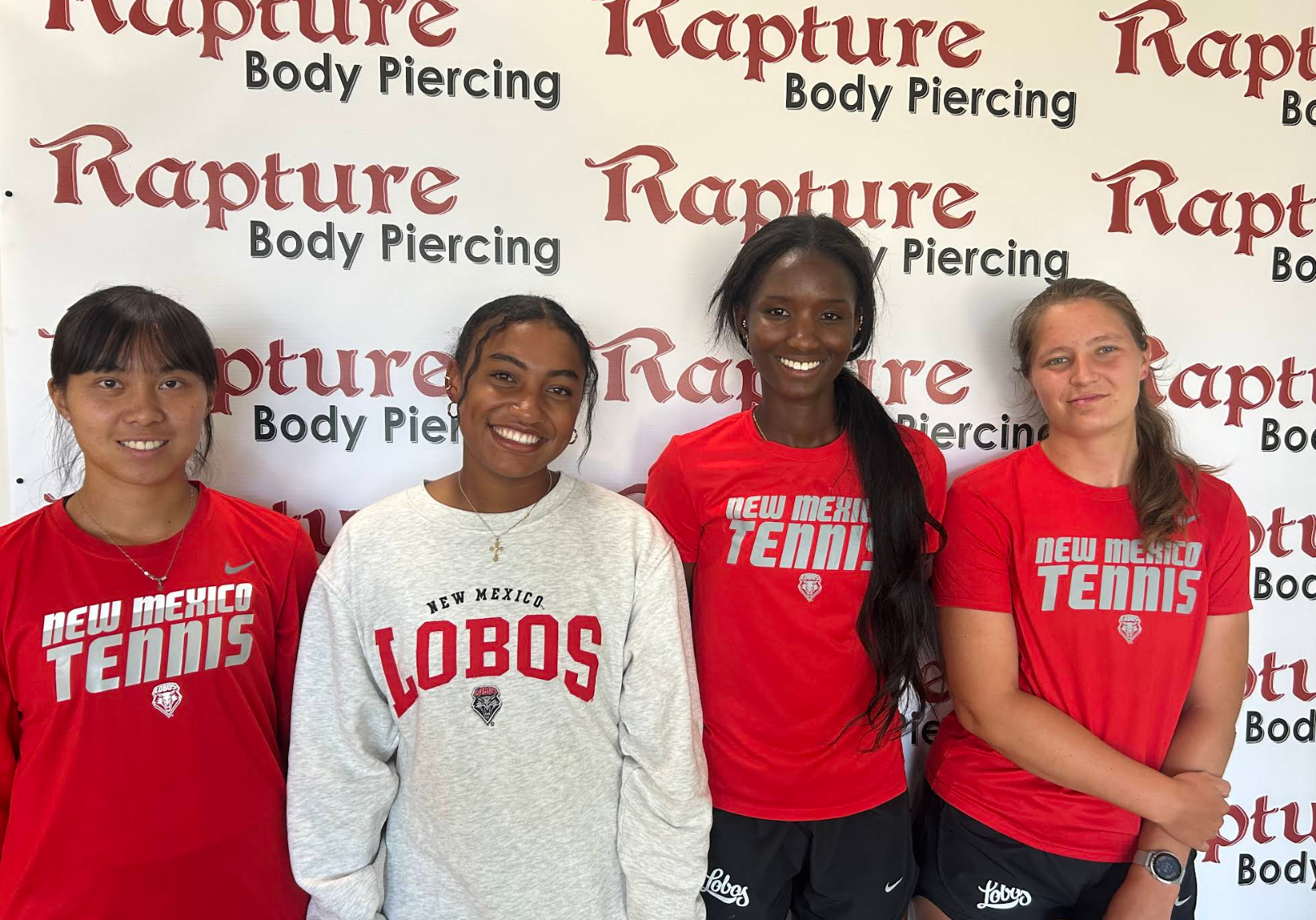 Four young women standing in front of a background with 'Rapture Body Piercing' printed repeatedly. They are wearing sports shirts, three are red and one is white, representing New Mexico Tennis and Lobos.
