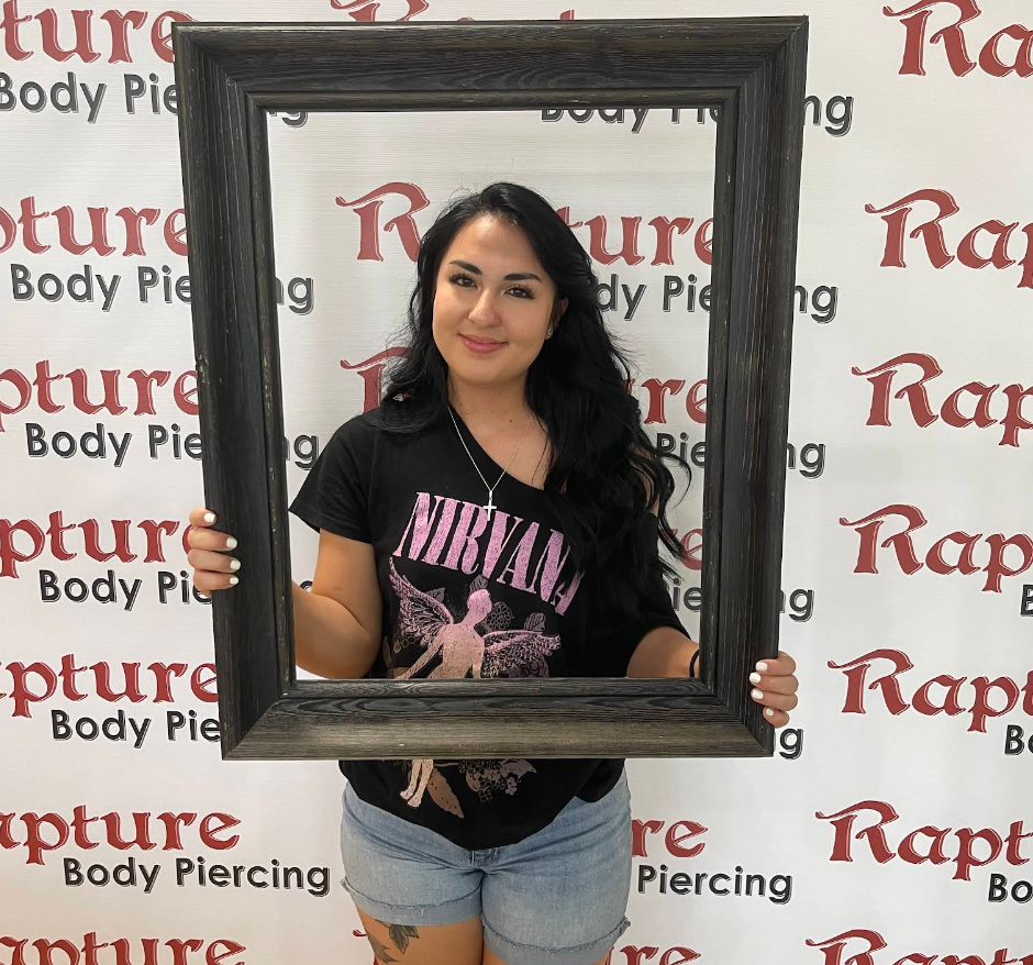 A young woman with long black hair holding a large dark wooden picture frame, standing in front of a background with repeated red and black text that says "Rapture Body Piercing." She is smiling at the camera, wearing a black Nirvana t-shirt and ligh
