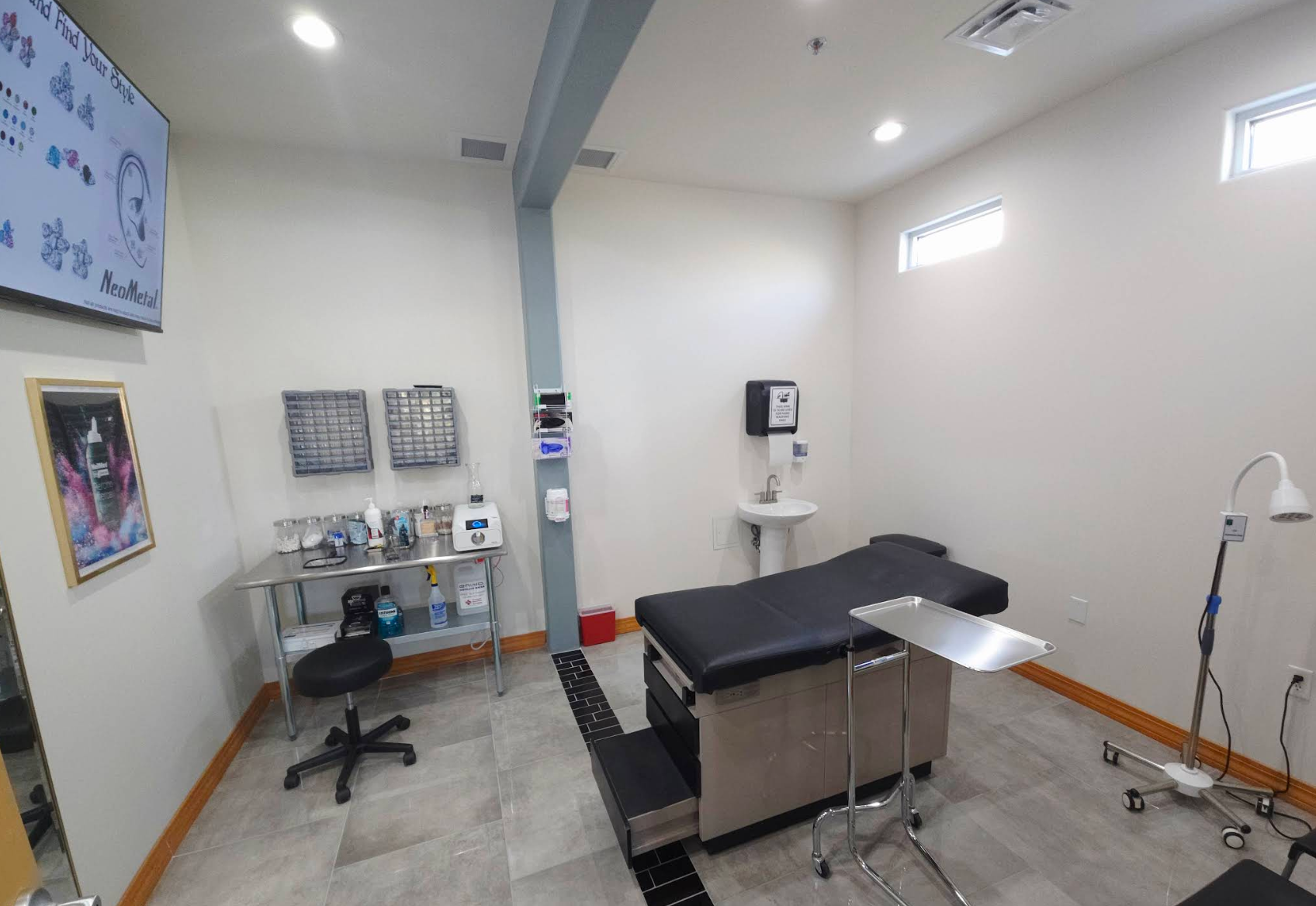 Medical examination room with a black examination table, medical supplies on a metal cart, wall-mounted sink, and medical equipment. The room has white walls, a small high window, and a floor with gray tiles.