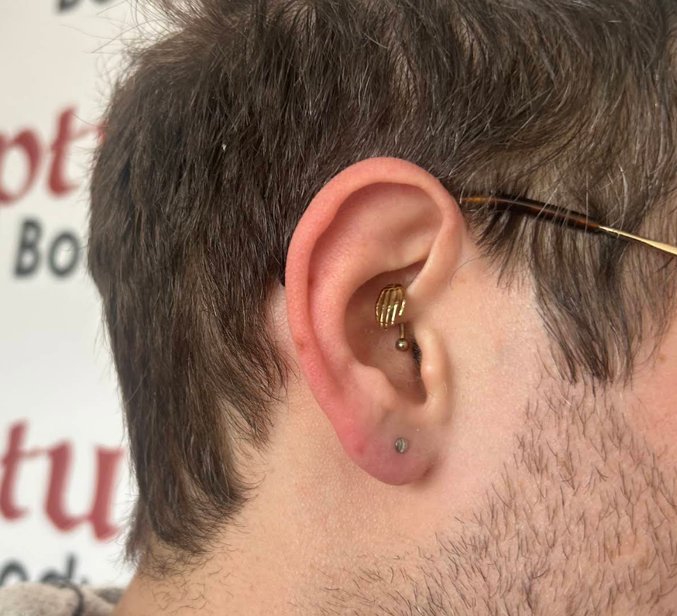 Close-up of a person's ear with three piercings: a cartilage piercing with a gold ring, a small earlobe piercing with a gem, and a second small earlobe piercing with a tiny gold ball earring.