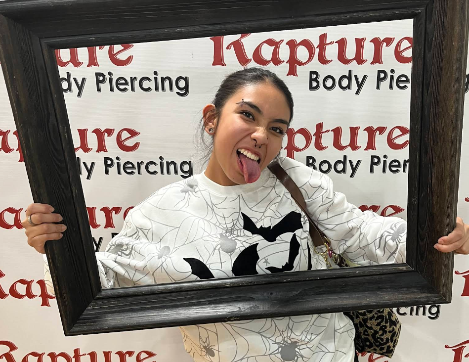 A young woman posing with a playful expression sticking her tongue out, holding a large empty picture frame, in front of a background that reads 'Capture Body Piercing' repeatedly.