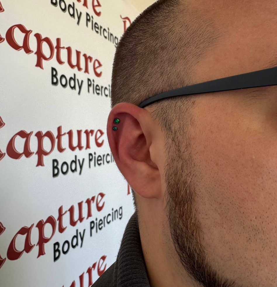 Close-up of a man's left ear with two green earrings, wearing glasses and a dark shirt, with a background featuring the words "Capture Body Piercing" repeated on a white wall.