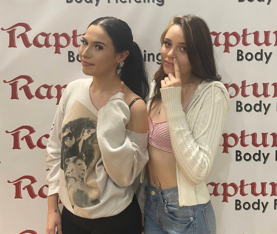 Two young women standing in front of a backdrop with red and black text that reads "Rapture Body Piercing." One woman has dark hair tied back, is wearing a graphic sweatshirt, and is smiling slightly. The other woman has brown hair, is wearing a whit