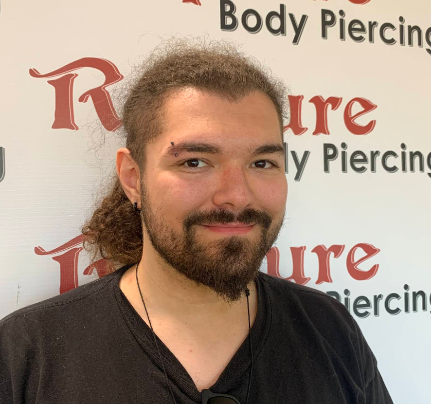 A man with a beard and curly hair, wearing a black shirt and earrings, standing in front of a wall with the words 'Body Piercing' and 'Pierure' visible.