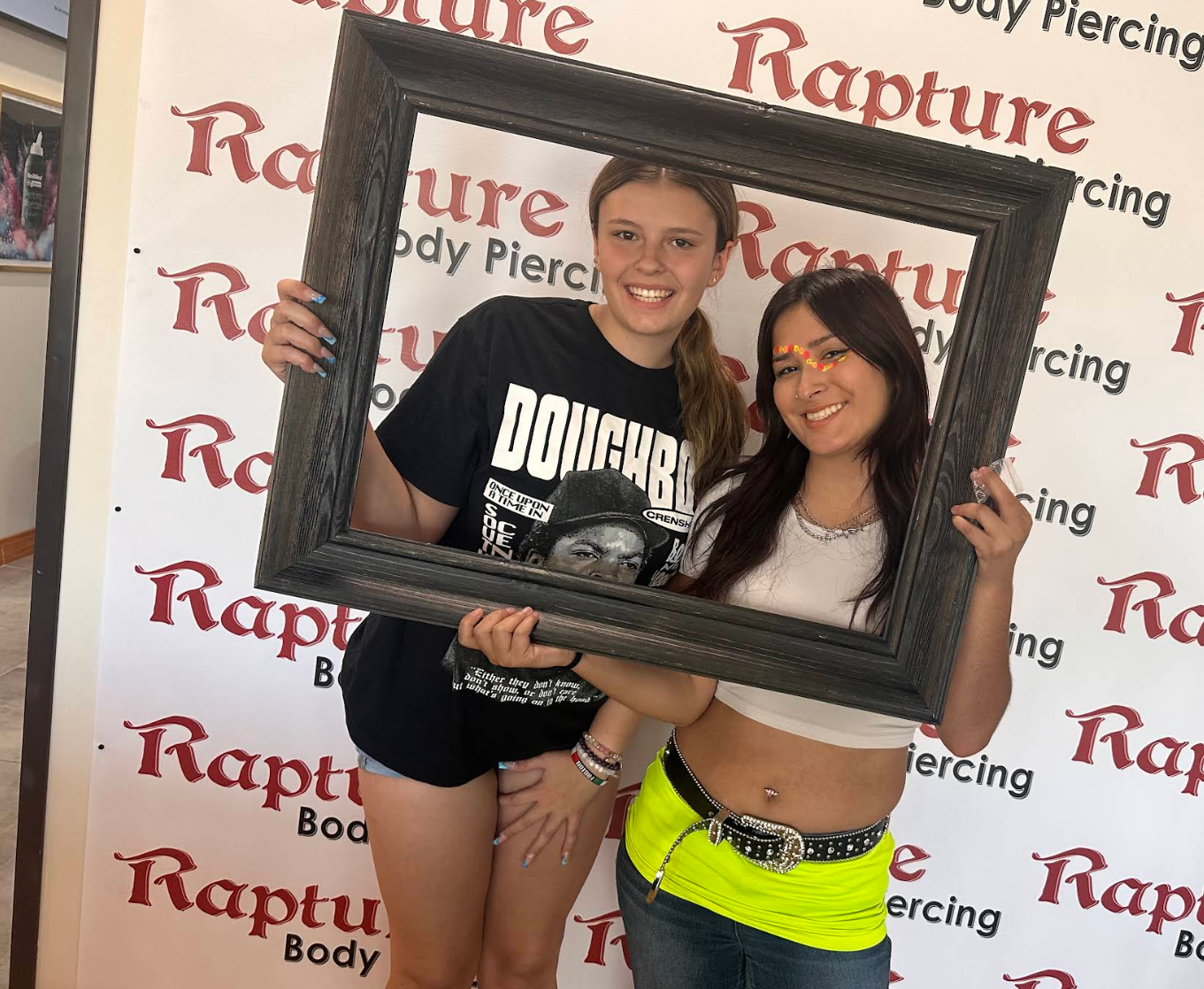 Two women, one in a black t-shirt and one in a white crop top, posing together behind a large empty picture frame. The woman on the right has face paint with rainbow colors and is wearing a neon yellow skirt with a black belt, while the woman on the 